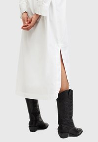 Abito camicia bianco con bottoni e spacchi sui lati, abbinato a stivali da cowboy neri in pelle con embossing floreale e tacco a blocco.