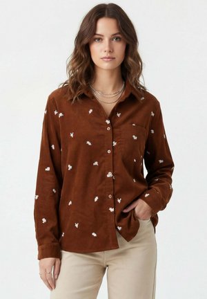 Femme aux cheveux bruns ondulés portant une chemise marron boutonnée avec des motifs floraux blancs, un pantalon beige et des colliers argentés superposés.