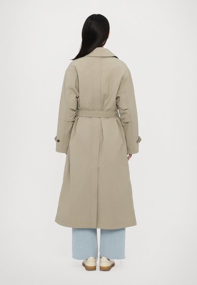Long beige trench coat with a belt, large collar, and buttoned cuffs, paired with light blue wide-leg jeans and cream shoes.