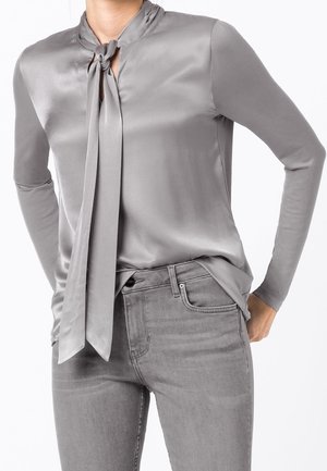 Szara satynowa bluzka z długimi rękawami i wiązaniem przy szyi, z wycięciem w kształcie otworu pod wiązaniem. W połączeniu z szarymi dżinsowymi spodniami.