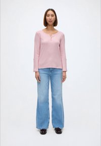 Απαλό ροζ μπλουζάκι μακρυμάνικο με λαιμόκοψη henley και τρία κουμπιά, συνδυασμένο με ανοιχτό μπλε παντελόνι με φαρδιές leg και μαύρα παπούτσια.