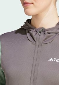 Pelēks zip-up džemperis ar teksturētu virsmu, kuram no vienas puses ir mīksts zaļš akcentu un kapuce ar auklu. Diskrēta zīmola nosaukuma uzraksts uz krūtīm.