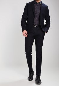 Costume bleu marine au design ajusté, avec une veste à un bouton et un pantalon à plastron. Accompagné d'une chemise à col foncé et de chaussures noires.