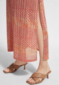 Robe maxi tricotée avec un motif en zigzag dans des nuances de rouge, orange et crème. Dispose d'une fente haute sur la cuisse. Chaussures à talons marron à enfiler.