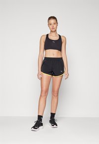Sostenedor deportivo negro con el logo de PUMA, combinado con pantalones cortos negros con ribetes amarillos y cordón. Zapatillas deportivas negras con acentos rosas.