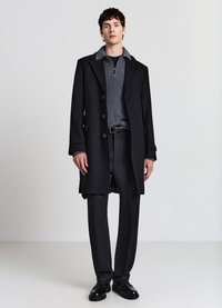 Cappotto nero con ampio colletto, maglione grigio con cerniera sotto, abbinato a pantaloni neri e scarpe nere. Tessuto liscio, vestibilità su misura.
