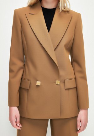 Femme portant un blazer croisé marron camel avec des boutons carrés dorés et un pantalon assorti, debout sur fond blanc.