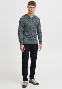 Mørkegrønn teksturert genser med V-hals, brukt med svarte slim-fit jeans og grå joggesko. Genseren har et melert mønster og ribbede mansjetter.