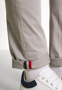 Pantalones beige con puños remangados que muestran una etiqueta roja y azul marino. Combinados con zapatillas blancas y calcetines blancos acanalados. Textura de tela suave.