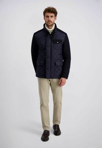 Mann, der eine marineblaue Steppjacke, einen cremigen Rollkragenpullover, beige Hosen und dunkelbraune Schuhe trägt, steht vor einem schlichten hellen Hintergrund.