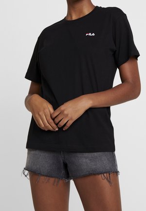 Osoba w czarnej koszulce z logo FILA oraz przetartych szarych szortach dżinsowych, z rękami złączonymi z przodu na jednolitym tle.