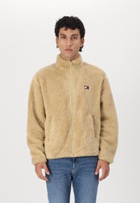 Hombre de pie vistiendo una chaqueta de forro polar beige con cremallera y jeans azules frente a un fondo blanco liso.