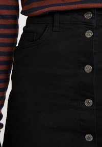 Černá džínová sukně s přímým střihem, s předním zapínáním na knoflíky a boční kapsou. Texturovaná látka se stříbrnými kovovými doplňky.