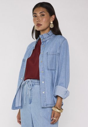 Donna che indossa giacca e pantaloni di jeans azzurro chiaro con top bordeaux, accessoriata con grandi orecchini, braccialetti e anelli, in piedi su uno sfondo semplice.