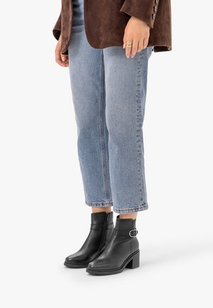 Personne portant un jean bleu clair, des bottines noires avec boucle et une veste marron, debout sur un fond blanc.