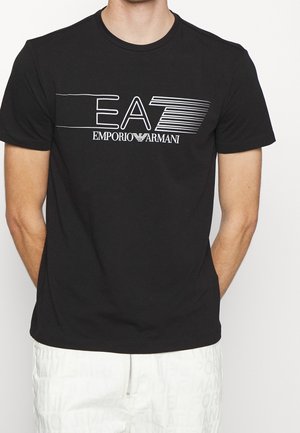 Homme portant un T-shirt noir avec le logo blanc "EA7 Emporio Armani" et un short blanc avec un motif de texte subtil, debout devant un fond blanc.