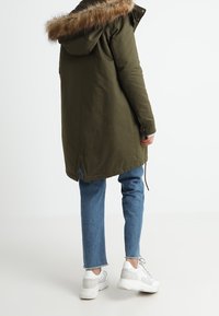 Parka vert olive avec une capuche doublée de fourrure amovible, présentant une coupe droite et une taille ajustable par un cordon, associée à un jean bleu clair et des baskets blanches.