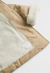 Beige Mantel mit einem weichen, cremefarbenen Kunstpelzkragen. Das Innere besteht aus hellbeigem Stoff mit einer feinen Textur. Enthält ein kleines Etikett an der Seite.
