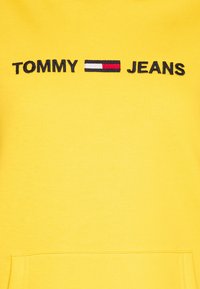 Geel sweatshirt met "TOMMY" en "JEANS" geborduurd in het zwart, met een rood en wit logo ertussen. Zacht materiaal met een voorzak.