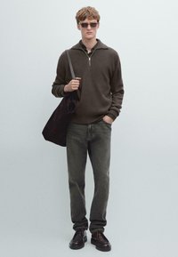 Brauner Strickpullover mit halbem Reißverschluss am Ausschnitt, kombiniert mit ausgewaschenen grauen Jeans und braunen Lederschuhen. Das Modell hält eine dunkle Tragetasche.