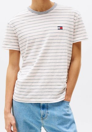 Osoba ubrana w biało-szarą koszulkę w paski z krótkim rękawem, szarym kołnierzykiem oraz jasnoniebieskie dżinsy, z ręką w kieszeni.