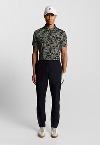 Man i vit keps, grön och svart mönstrad kortärmad skjorta, svarta byxor, vit golfhandske och vita sneakers, står mot en grå bakgrund.