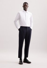 Camicia bianca a maniche lunghe con colletto e tasca anteriore, abbinata a pantaloni a pieghe blu scuro e scarpe stringate nere. Tessuto dalla superficie liscia.