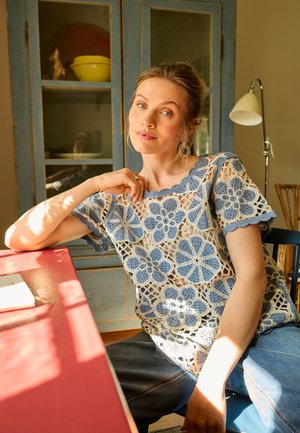Vrouw met licht haar die een blauw-wit gehaakt bloemen top draagt, zittend aan een rode tafel in een gezellige kamer met een kast en lamp.