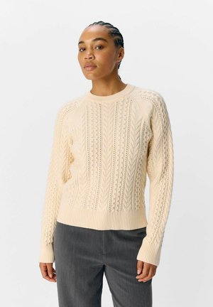 Vrouw met gevlochten haar in een crèmekleurige kabelgebreide trui en grijze broeken, staande tegen een witte achtergrond.