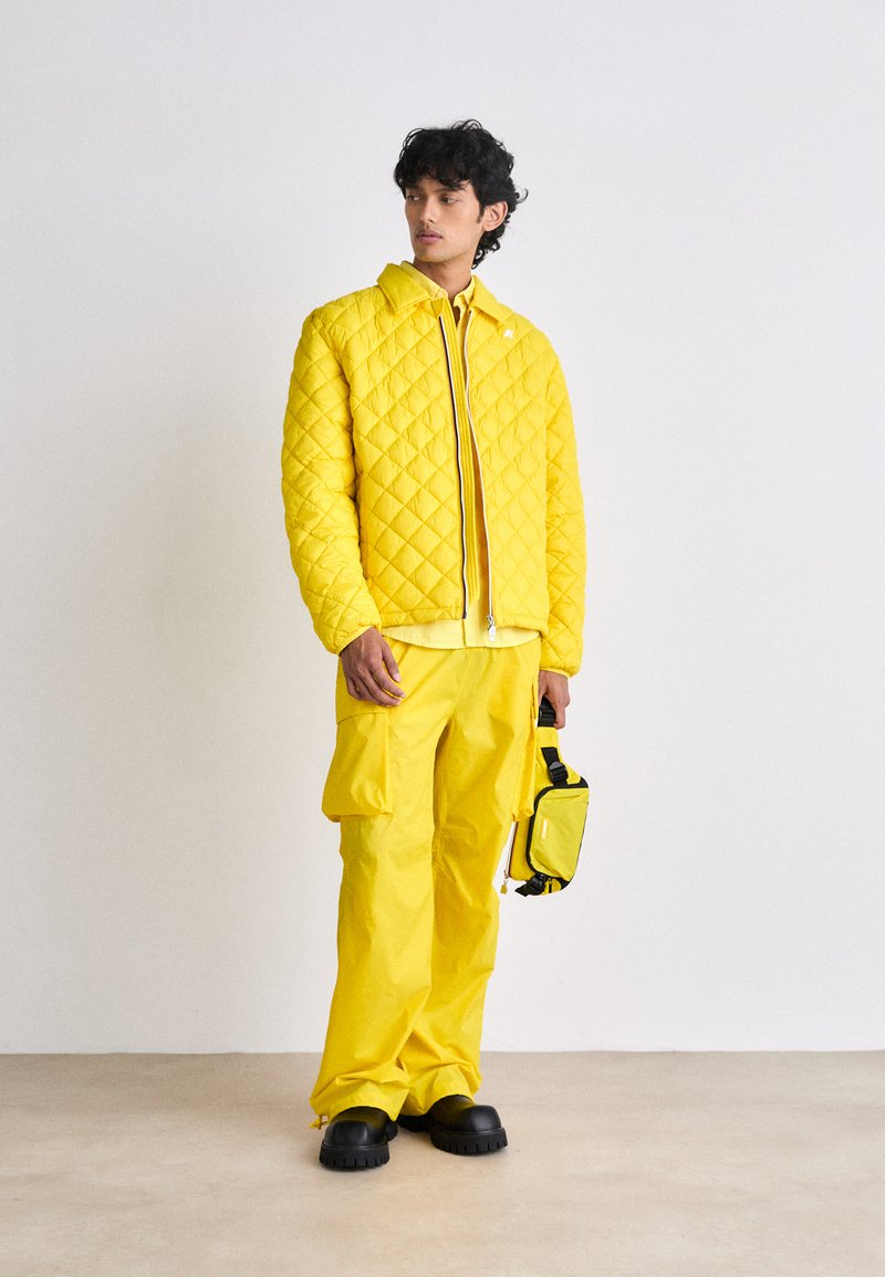 Casaco amarelo acolchoado com fecho de correr, combinado com calças cargo amarelas a condizer e uma pequena mala de tiracolo amarela. Sapatos pretos completam o visual.