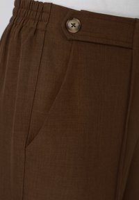 Pantalon marron sur mesure avec poche latérale et détail bouton sur la ceinture élastique, porté avec un haut blanc.