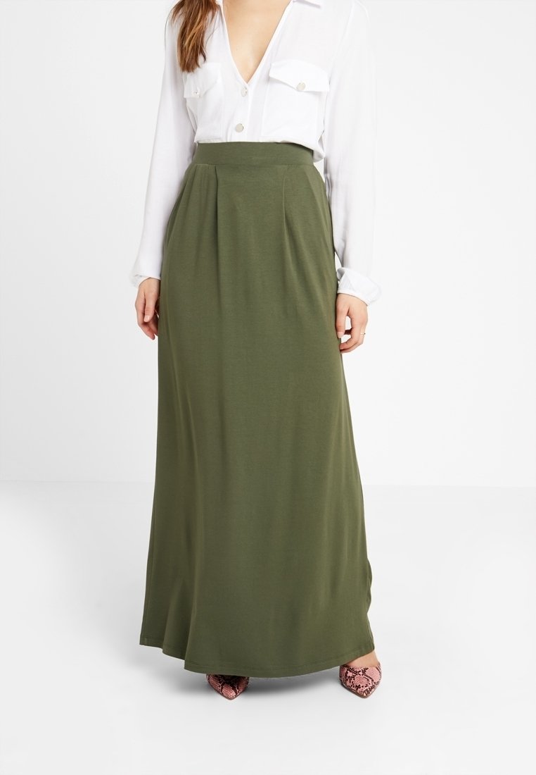 Jupe maxi vert olive à plis, en tissu doux. Associée à un chemisier blanc et des chaussures roses à motif. Texture lisse, longueur jusqu'à la cheville.