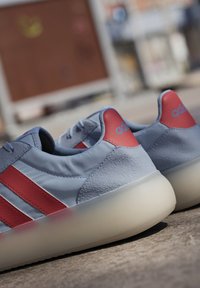 Plave tenisice izrađene od glatkih i teksturiranih materijala, s crvenim prugastim naglascima i prozirnom potplatom. Ikonski logo na peti.