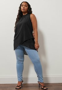 Top senza maniche nero con un collo alto e un orlo asimmetrico, abbinato a jeans skinny blu chiaro e scarpe con tacco nere a fasce. Texture liscia.