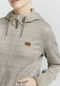Veste à capuche avec un motif géométrique beige et blanc, dotée d'une fermeture éclair intégrale et d'un cordon de serrage beige. Texture douce et détail de logo visibles.