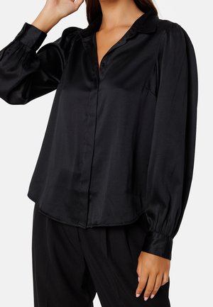 Femme portant un chemisier en satin noir à manches longues avec col et poignets boutonnés, associé à un pantalon noir, sur fond blanc.