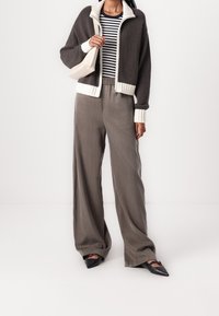 Szara kurtka zapinana na zamek z białym ściągaczem przy kołnierzu i mankietach, w połączeniu z luźnymi szarymi spodniami i czarnymi czubkowymi butami. Pod spodem widoczna paskowana bluzka.