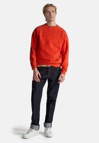 Oranžová fleece mikina s kulatým výstřihem, žebrovanými manžetami a lemem, kombinovaná s tmavými džínami a bílými teniskami s ohrnutými lemy.