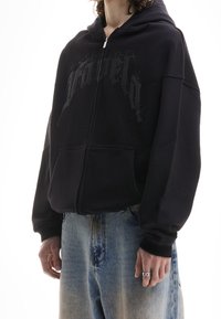 Personne portant un hoodie noir zippé avec de grandes poches avant et un jean bleu délavé, montrant une bague en chaîne en argent sur la main droite.