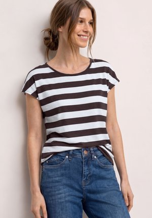 Kobieta uśmiechająca się, ubrana w czarno-biały krótki top w poziome paski z niebieskimi jeansami, stoi na tle jasnej ściany.