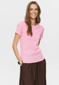 Tricou din bumbac roz, cu mâneci scurte și decolteu rotund. Purtat cu pantaloni maro, având o brățară elegantă din aur la încheietură.