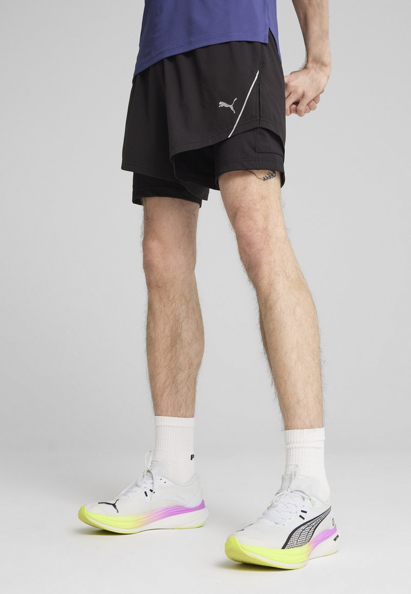 Sort atletiske shorts, lagdelt design, Puma-logo. Hvide sneakers med gule og pink accenter, tekstureret overflade, mønster på sålen. Hvite sokker.