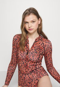 Långärmad baddräkt i orange med svart leopardmönster, med dragkedja och rund logotypdetalj på bröstet. Slät textur.