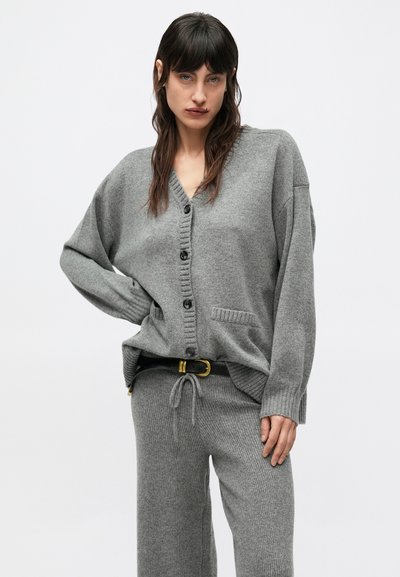Szary, oversize'owy kardigan z dekoltem w serek, guzikami z przodu i prążkowaną fakturą. Dobrany do pasujących szarych spodni i czarnego paska.