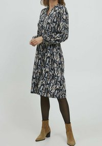 Robe à manches longues à motifs dans des teintes marine, beige et grise, avec un col à boutons et une jupe à volants, portée avec des bottines beiges.