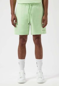 Pantalones cortos de algodón verde claro con cintura elástica y cordón, con un logo en el lateral. Combinados con calcetines blancos y zapatillas.