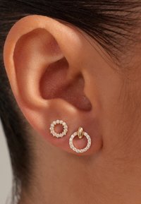 Boucles d'oreilles en or et diamants : l'une circulaire à surface lisse, l'autre texturée avec de petits diamants, portées sur une oreille teintée de peau.
