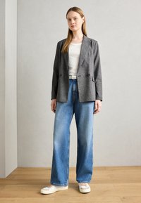 Blazer cinza com padrão sobre uma camisa branca, combinado com calças de ganga azuis folgadas e ténis brancos. Tecido texturizado; corte descontraído.