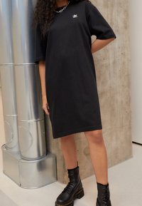 Černé bavlněné šaty ve volném střihu, s krátkými rukávy a malým bílým logem na hrudi, doplněné černými šněrovacími bojovými botami.