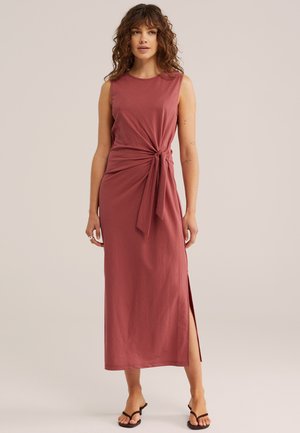 Žena kučeravými vlasmi nosiaca bezrukávové midi šaty v prašnej ružovej farbe s bočným rozparkom a zviazaným detailom v páse, stojaca v čiernych sandáloch.